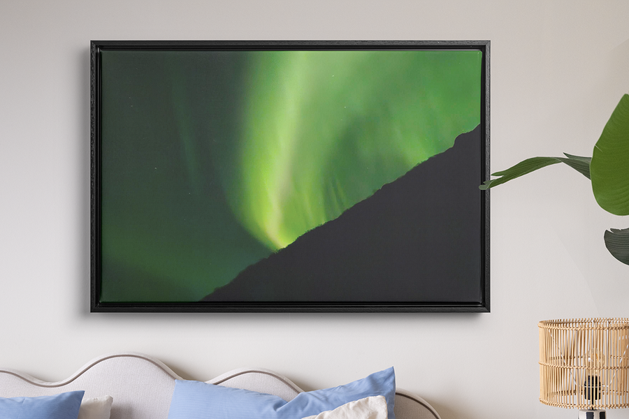 A framed print of very green northern lights hanging on a wall above a bed and a raffia lamp, illustrating how Photobox’s wall art turns northern lights photos into something worth keeping.