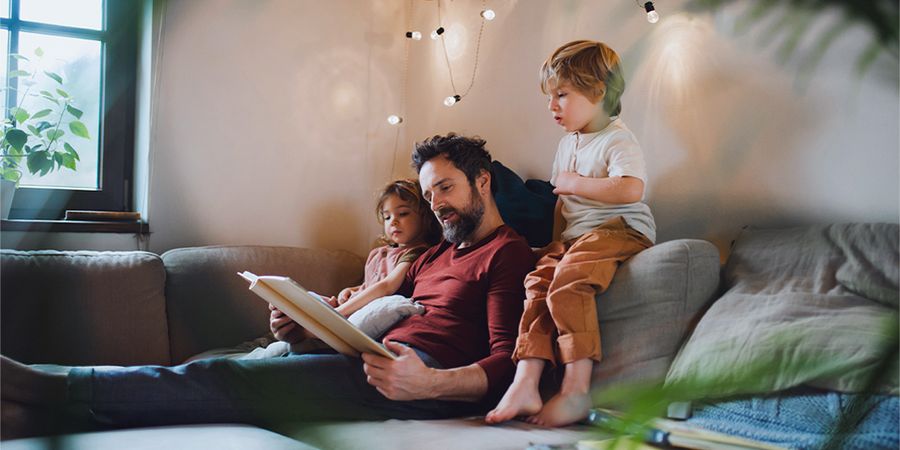 A dad reads a book to a young boy and girl on the sofa, with warm fairy lights glowing in front of them, introducing Photobox's cosy autumn hygge decor tips.