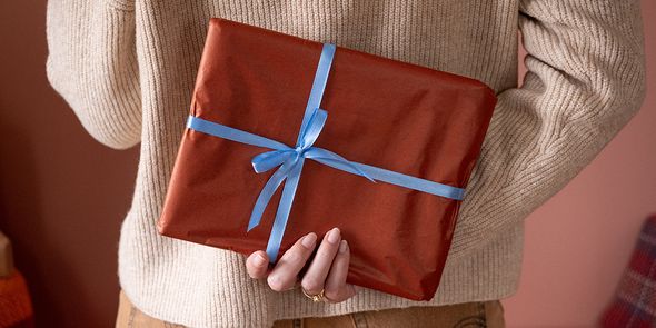 Woman holding a gift wrapped in brown paper with a blue ribbon behind her back, illustrating Photobox’s Christmas gift ideas checklist for a personalised photo gift.