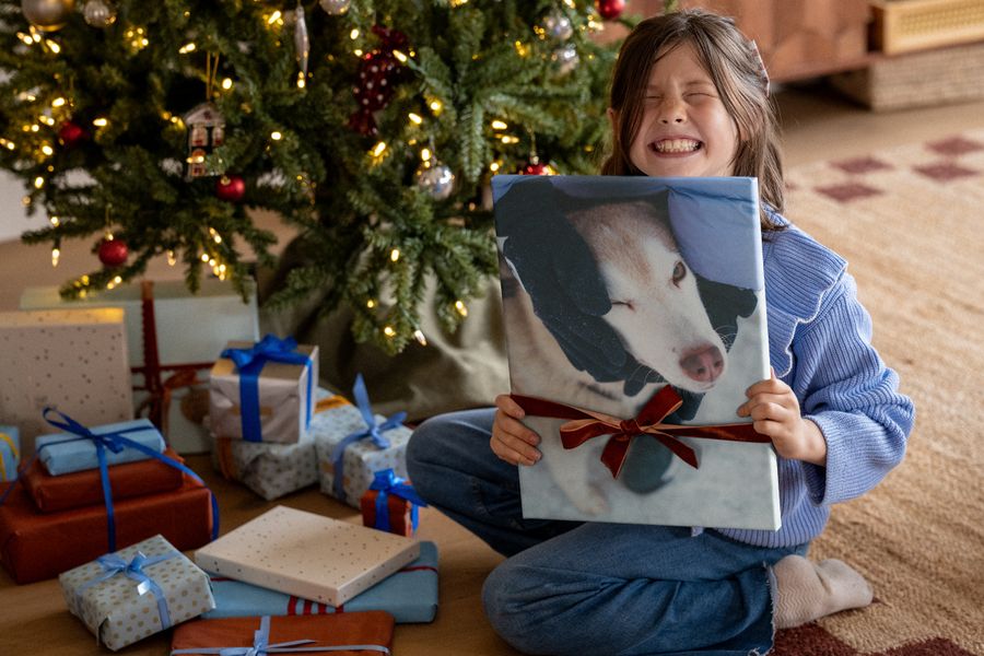 A laughing girl in a blue jumper holds up a canvas print of a dog with a red ribbon tied around it, in front of a Christmas tree, illustrating Photobox’s wall art for photos that deserve pride of place.