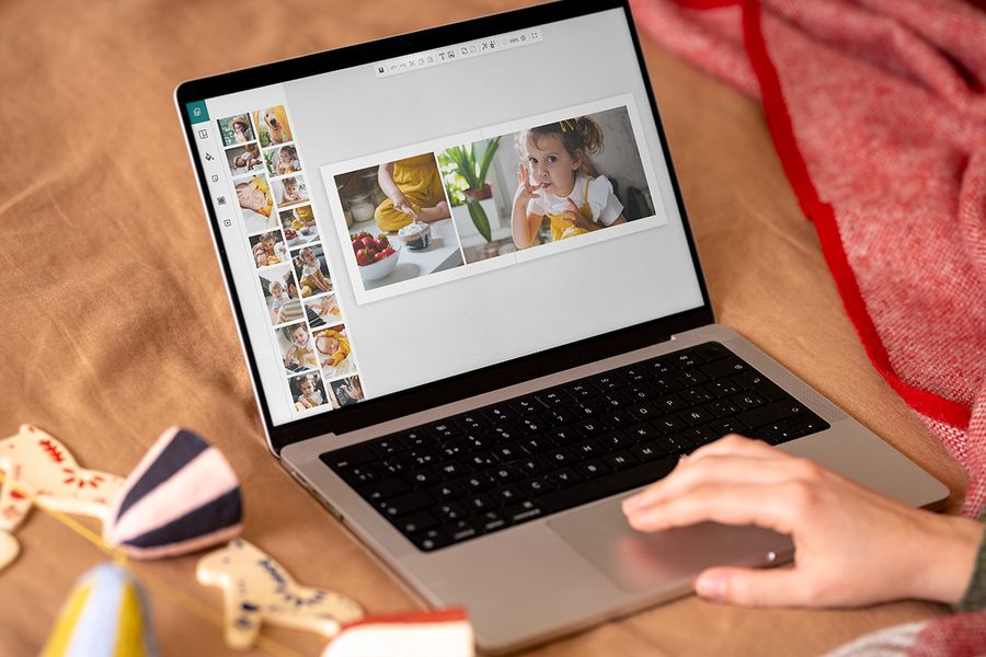 A hand on a laptop trackpad designs a photo book on screen, with a spread of a child and a bowl of strawberries, illustrating how to get creative online with Photobox’s photo book editor before Christmas.