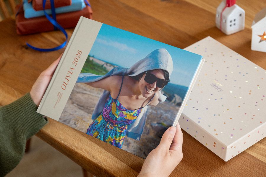 Two hands hold a photo book with 2026 on the cover, showing a smiling woman in sunglasses on a rocky coast, beside a confetti-patterned presentation box, illustrating Photobox’s personalised photo books.