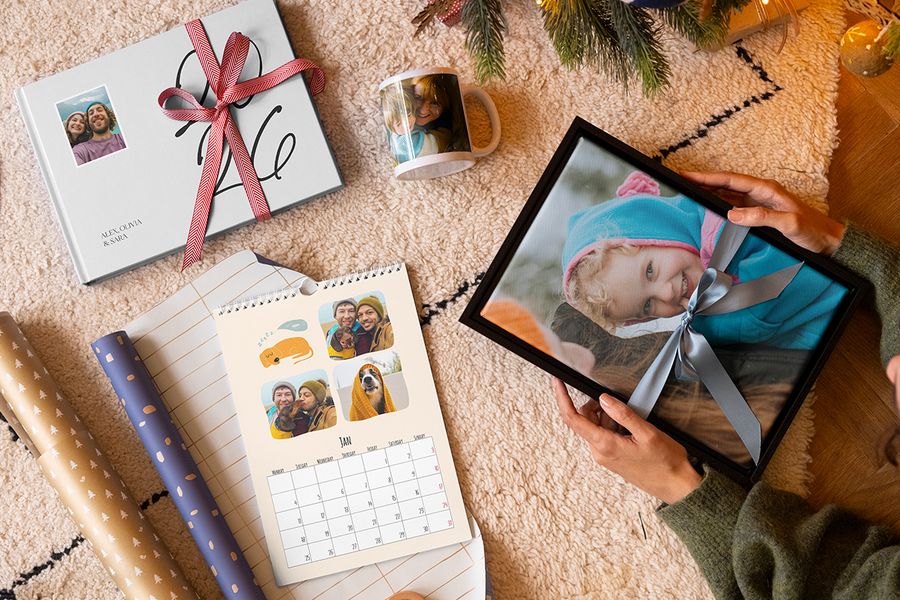Selection of Photobox personalised gifts, including wall art of a young girl with a blue ribbon around it, a mug with a mother and child, a personalised photo calendar and a 2026 photo book with a red ribbon around it, next to wrapping paper with Christmas tree branches in the background, illustrating Photobox’s tips on capturing the right memories for a personalised Christmas gift.
