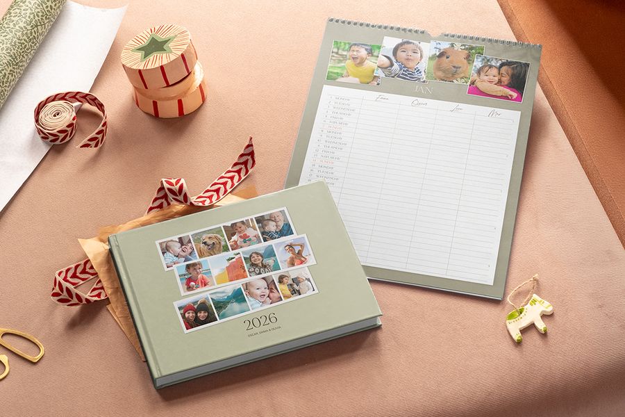 A sage-green 2026 photo book sits beside a family wall planner with photos along the top and columns for each family member, illustrating Photobox’s shared family planner for Black Friday Christmas gifts.