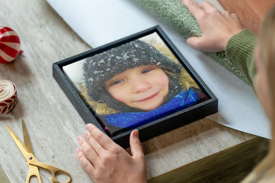 Framed canvas of a young girl in winter clothes in the snow, with a woman’s hands ready to wrap it beside wrapping paper, scissors and ribbon, illustrating Photobox’s tips on choosing a personalised photo gift to suit someone’s style.