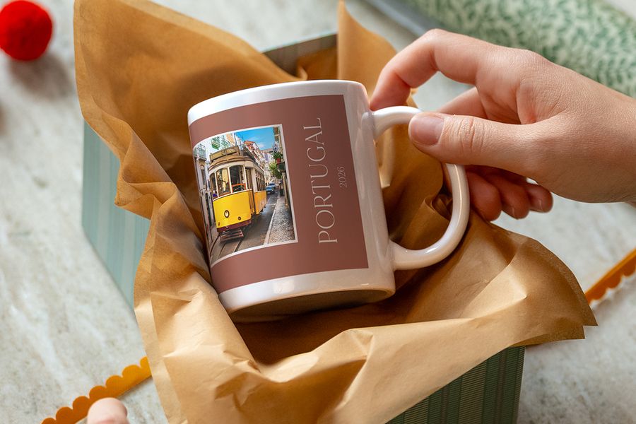 A hand lifts a mug wrapped in kraft tissue paper, printed with a yellow tram and the words ‘Portugal 2026’, illustrating Photobox’s custom mugs as a clue for an experience gift.