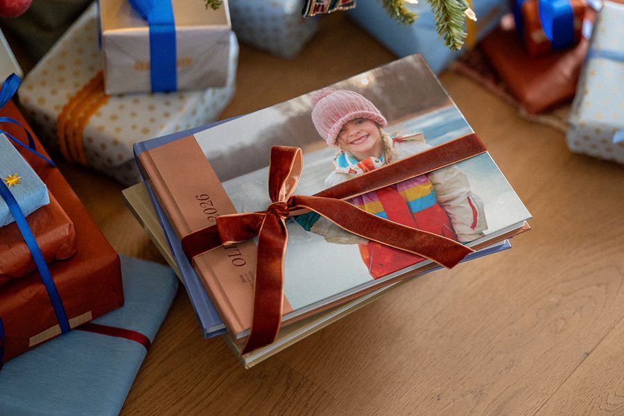A stack of photo books tied with a velvet ribbon, the top cover showing a smiling girl in a pink knitted hat and 2026 on the spine, placed among wrapped Christmas presents, illustrating Photobox’s photo book of the year.
