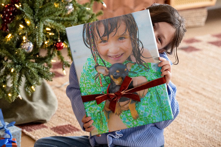 Little girl under the Christmas tree holding a large photo book with a velvet ribbon around it and a photo of herself with a chihuahua on the cover, illustrating Photobox’s checklist for a personalised Christmas photo gift.