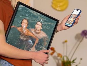 A woman holds her phone showing the Photobox editor while carrying holiday wall art under her arm, featuring two males having fun in a pool, inspiring ways to turn holiday photos into stunning wall art.