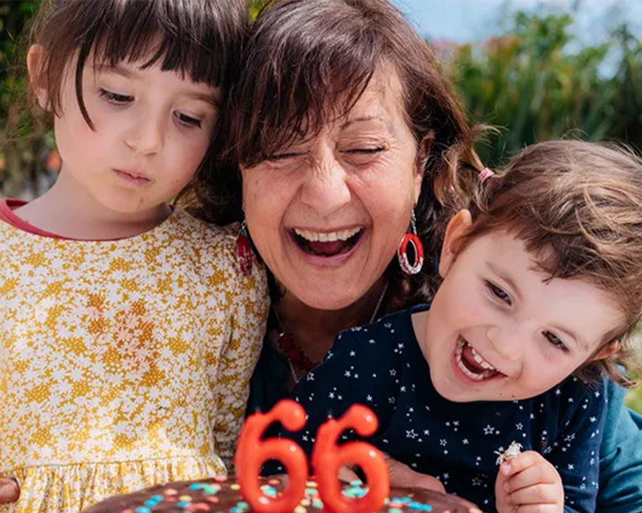 A grandmother celebrates her 66th birthday with her two grandchildren beside her, about to blow out the candles on her cake, inspiring birthday gift ideas from the Photobox ultimate birthday gift guide.